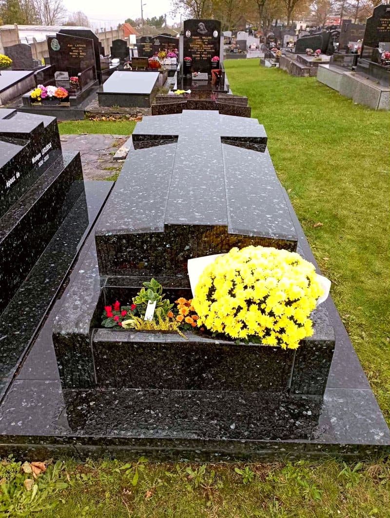Une tombe en granit noir surmontée d’une croix, des chrysanthèmes jaunes.