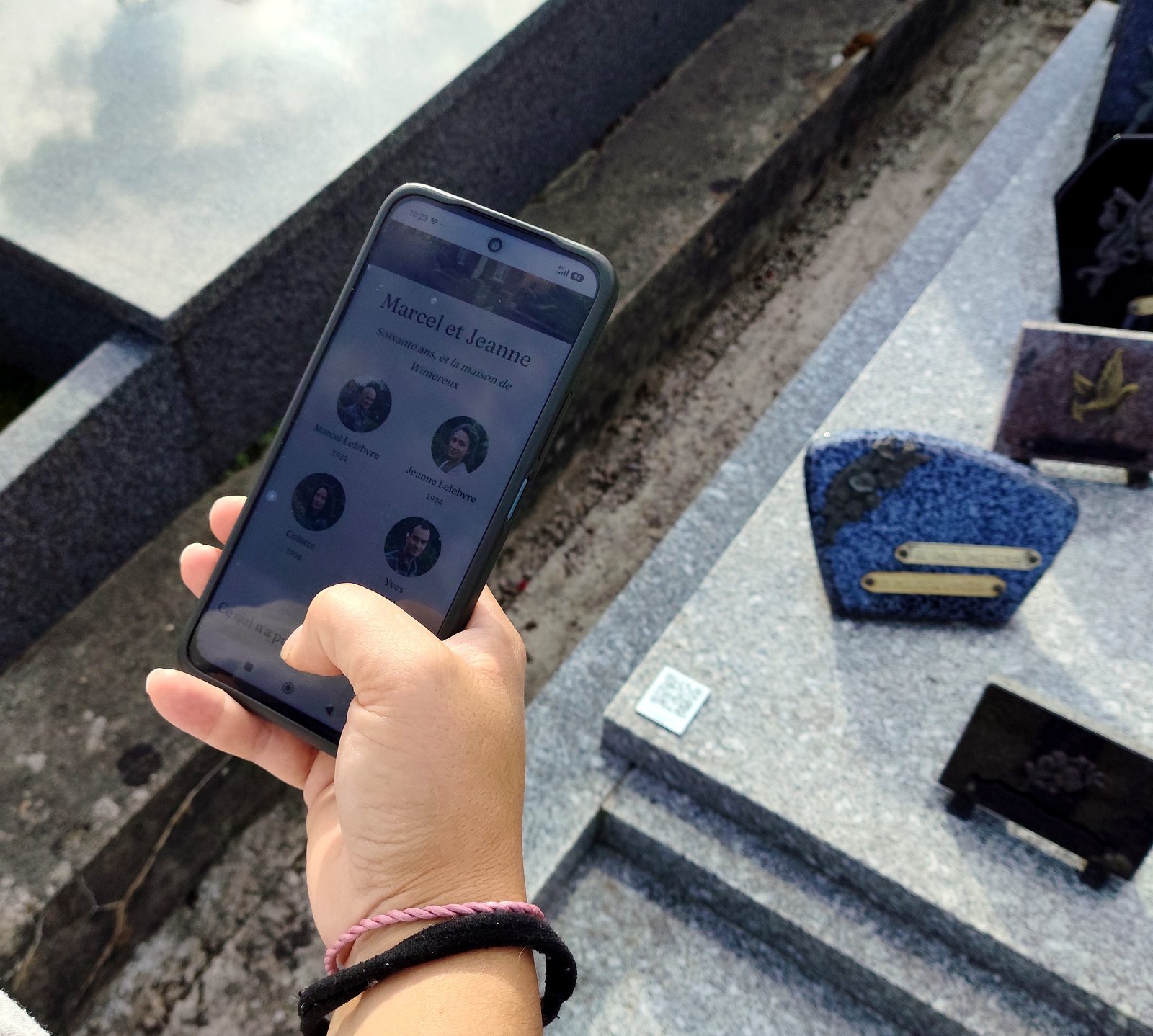 Une main tient un téléphone devant une sépulture : la page de l'Histoire est ouverte à l'écran, et la petite plaque au code est posée sur le granit à droite.